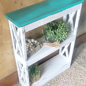 May include: A narrow, two-tiered wooden shelf unit with a distressed white finish and a solid turquoise top. The sides feature a white crisscross X-pattern. The shelves hold a small green plant, a wooden bowl with white beads, and a small gold pot.