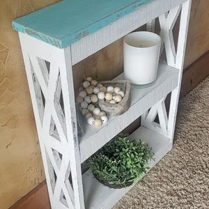 May include: A narrow white wooden shelf unit with a distressed finish and a turquoise top surface. The sides feature an X-pattern design. The middle shelf holds a white candle and a string of wooden beads, while the bottom shelf holds a green plant.
