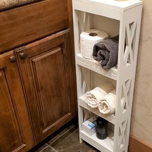 May include: A tall, narrow white wooden storage shelf with three open tiers and X-shaped side panels. The unit holds a roll of toilet paper, a dark gray towel, two rolled white towels, a small box, and a black cylindrical container.