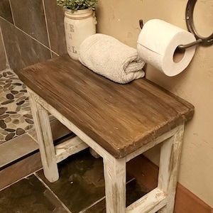May include: A wooden bench with a dark brown stained top and distressed white painted legs. On the bench sits a white towel and a white jar containing a green plant. A roll of white toilet paper hangs from a metal horseshoe wall mount.