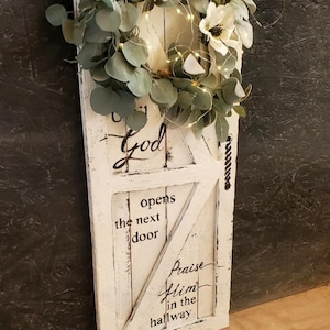 May include: A distressed white wooden wall hanging shaped like a door, adorned with a floral and eucalyptus garland and fairy lights. The text reads "God opens the next door, Praise Him in the hallway."