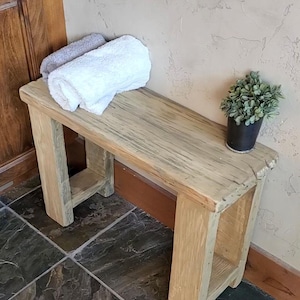 May include: A rectangular wooden bench with a light brown, natural wood grain finish and thick, sturdy legs. On the bench, there are two folded towels, one white and one grey, and a small potted green plant in a dark grey container.