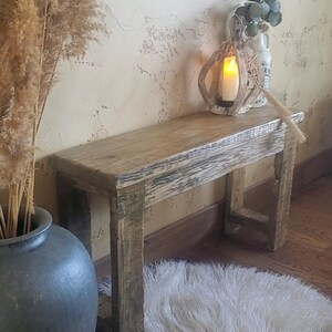 May include: A rectangular wooden bench with a weathered, light brown finish and visible wood grain. The bench features four sturdy legs and a flat top surface, constructed from reclaimed-style timber with a rough, textured appearance.