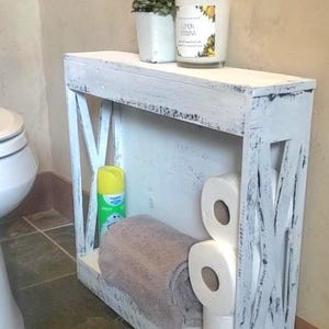 May include: A white distressed wooden storage shelf with X-shaped side panels. The shelf holds a grey towel, three rolls of toilet paper, a yellow spray bottle, a small potted plant, and a candle with a label reading Lemon Verbena.