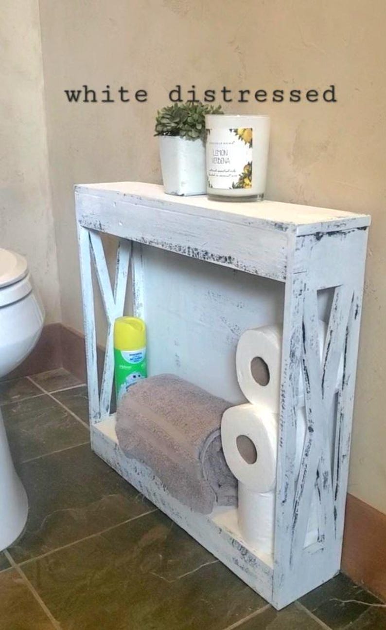 May include: A white distressed wooden storage shelf with X-shaped side panels. The shelf holds a grey towel, three rolls of toilet paper, a yellow spray bottle, a small potted plant, and a candle with a label reading Lemon Verbena.