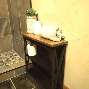 May include: A narrow wooden bathroom storage shelf with a dark charcoal finish and a natural wood top. The unit features an X-shaped side frame design, two open shelves, and holds a white toilet paper roll, a white soap dispenser, and a towel.