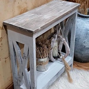 May include: A rectangular console table with a distressed white finish and a natural wood-toned top. The table features X-shaped side supports and a lower shelf. On the shelf sits a metal tin containing dried grasses and a decorative wooden object.