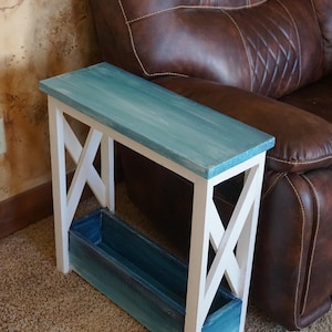 May include: A wooden side table with a rectangular top and a built-in storage box. The top is painted in shades of blue, while the frame is white. The table has an X-shaped design on the sides. The table is 30 inches long.