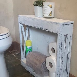 May include: A narrow, rectangular white wooden shelf with a distressed finish and X-shaped side supports. The shelf holds a small potted plant, a Lemon Verbena candle, a yellow aerosol spray can, a folded gray towel, and three rolls of toilet paper.