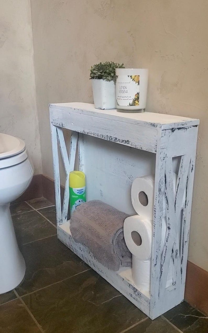 May include: A narrow, rectangular white wooden shelf with a distressed finish and X-shaped side supports. The shelf holds a small potted plant, a Lemon Verbena candle, a yellow aerosol spray can, a folded gray towel, and three rolls of toilet paper.