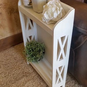 May include: A narrow white wooden two-tier shelf featuring side panels with cutout X-shaped designs. The top shelf holds a white candle jar and a white flower, while the bottom shelf contains a small green artificial plant.
