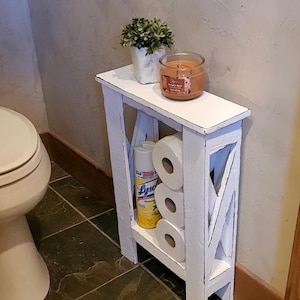 May include: A white, distressed-look wooden storage shelf with an X-shaped design on the sides. It holds three rolls of toilet paper, a Lysol spray bottle, a small potted plant, and a candle in a glass jar.