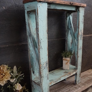 May include: A rectangular wooden console table featuring a distressed light blue paint finish with visible dark wood grain. The structure includes four vertical legs, a flat top surface, and a lower shelf with a small square planter placed on it.
