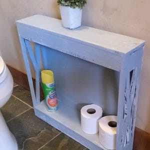 May include: A narrow, rectangular, gray distressed wooden shelf unit with an X-pattern side design. The top surface holds a small white pot with a green plant. The bottom shelf contains a yellow aerosol spray can and two rolls of white toilet paper.