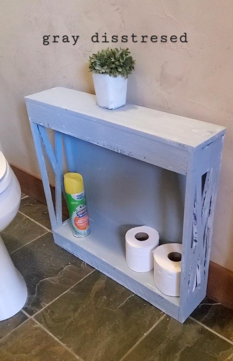 May include: A narrow, rectangular, gray distressed wooden shelf unit with an X-pattern side design. The top surface holds a small white pot with a green plant. The bottom shelf contains a yellow aerosol spray can and two rolls of white toilet paper.
