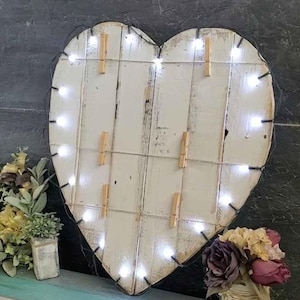 May include: A heart-shaped wall display made of white-painted wooden planks with a distressed finish. The piece features thin wire strands strung across the front, accented with small wooden clothespins and a string of bright white LED lights.