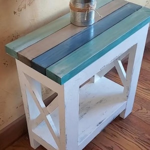 May include: A rectangular side table with a white distressed wooden frame and an X-shaped side support. The tabletop consists of horizontal planks painted in shades of teal, blue, and grey. A silver metal tin filled with dried grass sits on top.