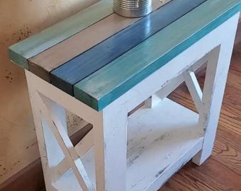 May include: A rectangular side table with a white distressed wooden frame and an X-shaped side support. The tabletop consists of horizontal planks painted in shades of teal, blue, and grey. A silver metal tin filled with dried grass sits on top.