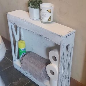 May include: A narrow, rectangular white wooden shelf with a distressed finish and X-shaped side panels. The shelf holds a gray towel, three rolls of toilet paper, a yellow spray bottle, a small potted green plant, and a candle labeled Lemon Verbena.