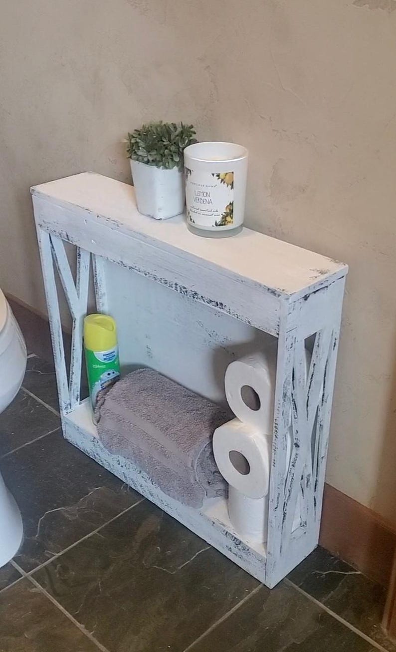 May include: A narrow, rectangular white wooden shelf with a distressed finish and X-shaped side panels. The shelf holds a gray towel, three rolls of toilet paper, a yellow spray bottle, a small potted green plant, and a candle labeled Lemon Verbena.