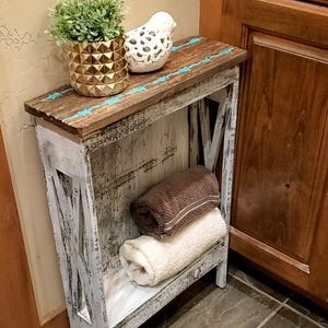 May include: A narrow wooden console table with a distressed white finish and an X-shaped side design. The top surface is dark brown wood with a turquoise inlay. Two rolled towels, one brown and one cream, sit on the lower shelf of the table.