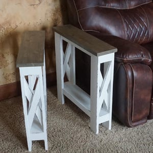 May include: Two white wooden side tables with gray-brown tops. The tables have an X-shaped design on the sides and a lower shelf. The tables are placed on a carpeted floor, with a brown leather recliner in the background.