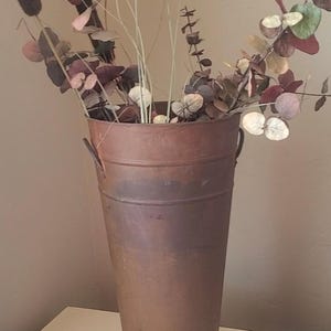 May include: A tall, cylindrical, rusted metal bucket with two small side handles, containing a decorative arrangement of dried eucalyptus branches with muted purple and green leaves and thin, light-colored ornamental grass stalks.