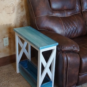 May include: A narrow, wooden side table with a rectangular top and a built-in planter box. The top is painted in shades of blue, while the frame is white. The table is designed with an X-shaped support structure. The table is next to a brown leather recliner.