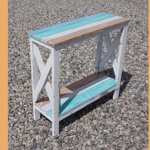 May include: A small, white wooden side table with a distressed finish. The tabletop features horizontal stripes of blue, white, and brown. The table has an open shelf and X-shaped supports on the sides.