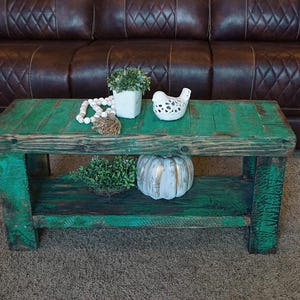 May include: A teal-painted wooden coffee table with a lower shelf. The table is decorated with a white bird figurine, a small white pot with greenery, a string of white beads, and a decorative pumpkin. A brown leather sofa is in the background.