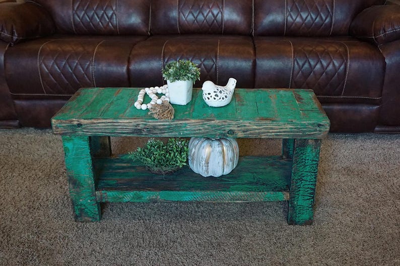 May include: A teal-painted wooden coffee table with a lower shelf. The table is decorated with a white bird figurine, a small white pot with greenery, a string of white beads, and a decorative pumpkin. A brown leather sofa is in the background.