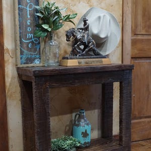 May include: A rustic wooden side table with a bronze sculpture of a cowboy on a horse. A white cowboy hat rests on the wall behind the sculpture. A turquoise jug with a cowboy silhouette and a welcome sign are also on the table.