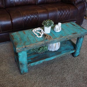 May include: A rustic, turquoise-painted wooden coffee table with a lower shelf. The table is adorned with decorative items, including a small potted plant, a string of white beads, and a decorative bird figurine. The table is placed on a brown carpet.
