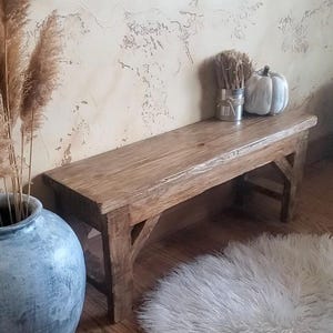 May include: A rectangular wooden bench with a natural brown finish, featuring four sturdy legs and angled support braces connecting the legs to the seat. The bench has a visible wood grain texture and a smooth, flat top surface for seating.