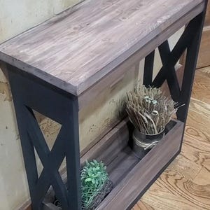 May include: A narrow console table featuring a rectangular top with a weathered brown wood finish and a black frame with X-shaped side supports. The base includes a lower shelf containing a small green plant and a metal container with dried grass.
