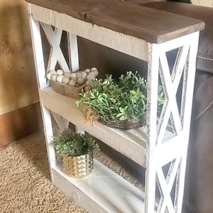 May include: A narrow wooden side table with a dark brown top and a distressed white frame featuring X-shaped side panels. The table has three open shelves holding a wooden bowl with light-colored beads, a green plant, and a gold textured pot.