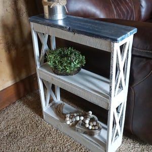 May include: A narrow wooden shelf unit with three tiers, featuring a distressed white finish on the frame and a dark gray top surface. The sides have a decorative X-pattern design. The unit holds a metal tin, a small green plant, and beads.