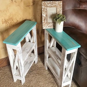 May include: Two rustic, white wooden side tables with turquoise tops and distressed finishes. Each table has an X-shaped side design and a lower shelf. A framed picture and a small plant sit on one table.