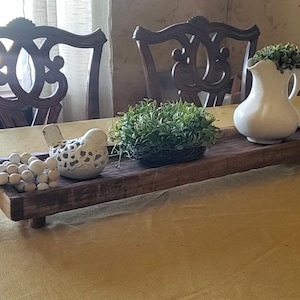 May include: A rustic wooden tray with decorative items. It features a white ceramic bird, a small white pitcher with greenery, a green plant ball, and a strand of wooden beads. The tray is on short legs and has a jute tassel.
