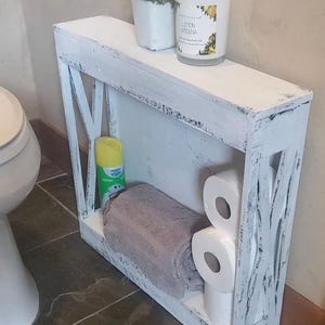 May include: A narrow, rectangular white wooden shelf with a distressed finish and X-shaped side supports. The shelf features a top surface holding a small potted plant and a candle, with a lower level containing a folded gray towel and toilet paper.