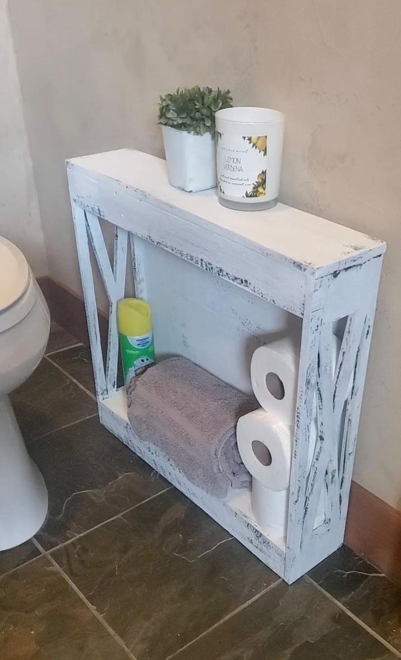 May include: A narrow, rectangular white wooden shelf with a distressed finish and X-shaped side supports. The shelf features a top surface holding a small potted plant and a candle, with a lower level containing a folded gray towel and toilet paper.