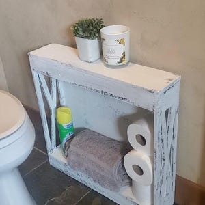May include: A narrow, rectangular white wooden shelf with a distressed finish and X-shaped side supports. The shelf holds a small potted plant, a Lemon Verbena candle, a yellow aerosol can, a folded gray towel, and two rolls of white toilet paper.