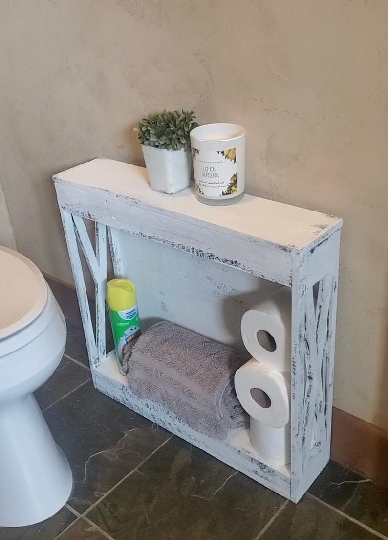 May include: A narrow, rectangular white wooden shelf with a distressed finish and X-shaped side supports. The shelf holds a small potted plant, a Lemon Verbena candle, a yellow aerosol can, a folded gray towel, and two rolls of white toilet paper.
