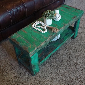 May include: A distressed, rectangular coffee table painted in a vibrant green. The table features a lower shelf and is accessorized with a white beaded garland, a small potted plant, and a decorative white ceramic piece. The table is made of wood.