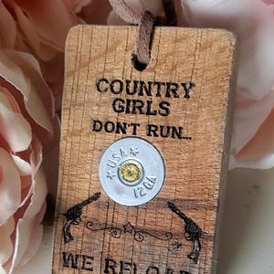 May include: A rectangular wooden tag with a brown leather cord, featuring the black text "COUNTRY GIRLS DON'T RUN... WE RELOAD". A silver 12 GA shotgun shell casing with "USA" stamped on it is centered, flanked by two black revolver illustrations.