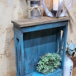 May include: A narrow wooden console table with a distressed teal blue finish and a dark brown top. The table features X-shaped side supports and a single open shelf. The surface holds a metal tin with dried grass and a white candle in a frame.