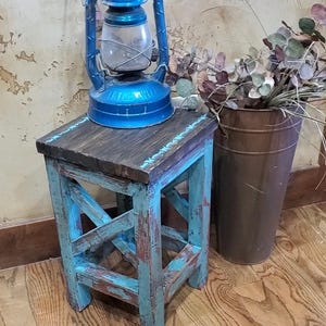 May include: A square wooden stool with a dark brown top and distressed turquoise blue legs featuring an X-frame design. A bright blue metal oil lantern with a clear glass globe sits on the stool, and a tall brown metal bucket holds dried foliage.