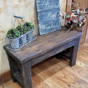 May include: A dark brown wooden bench with a rustic finish. On top sits a rectangular wire basket containing three small potted green plants and a gray wooden sign with white text that reads: Life doesn't have to be Perfect to be Wonderful.