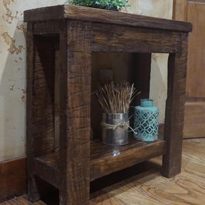 May include: A rustic, dark brown wooden side table with a lower shelf. The table has a rough-hewn texture. The top shelf holds a small green plant. The lower shelf displays a tin can with twine and dried grass, and a turquoise decorative lantern.