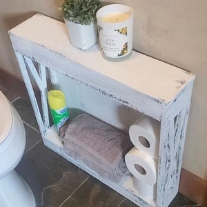 May include: A narrow, white, distressed-finish wooden shelf unit with X-shaped side supports. The top surface holds a small potted green plant and a Lemon Verbena candle. The lower shelf contains a folded gray towel, two toilet paper rolls, and a yellow spray bottle.